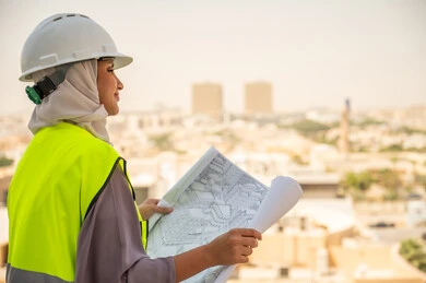  مشروع بناء ، خلفية عمارات وأبنية، صورة جانبية لمهندسة معمارية عربية سعودية خليجية تحمل بيدها مخطط البناء وتتأمل معالم المدينة ، مهندسة انشاءات محجبة ترتدي بدلة السلامة في العمل، لباس رسمي للعمل ، خوذة العمل, مراجعة المخططات الهندسية وتدقيقها