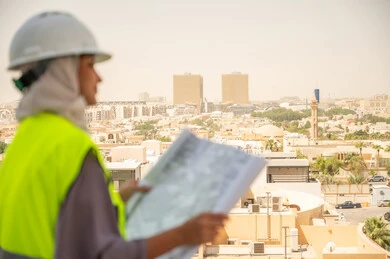  مشروع بناء ، خلفية عمارات وأبنية، صورة جانبية لمهندسة معمارية عربية سعودية خليجية تحمل بيدها مخطط البناء وتتأمل معالم المدينة ، مهندسة انشاءات محجبة ترتدي بدلة السلامة في العمل، لباس رسمي للعمل ، خوذة العمل, مراجعة المخططات الهندسية وتدقيقها