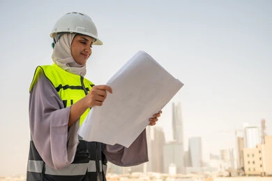 Side view of a Saudi Arabian Gulf female architect holding a building plan, a veiled female construction engineer wearing a safety suit, formal work clothes, work helmet, reviewing and checking engineering plans, construction project, background of buildings and structures