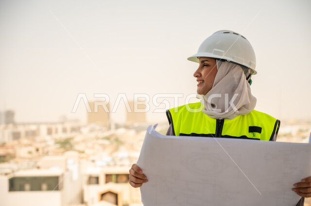  لباس رسمي للعمل ، خوذة العمل, مهندسة معمارية عربية سعودية خليجية تحمل مخطط البناء، مهندسة انشاءات محجبة ترتدي بدلة السلامة في العمل، تطور القطاع الهندسي في المملكة، خلفية أبراج وناطحات سحاب، مراجعة المخططات الهندسية وتدقيقها، مهنن ووظائف نسائية سعودية