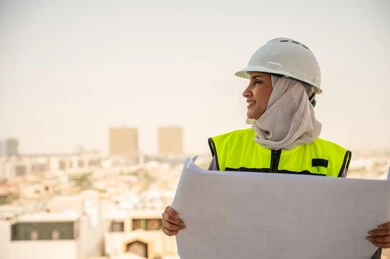 Formal work dress, work helmet, Saudi Arabian Gulf female architect holding a building plan, veiled female construction engineer wearing a safety suit at work, development of the engineering sector in the Kingdom, background of towers and skyscrapers, reviewing and auditing engineering plans, Saudi women's professions and jobs