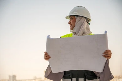  لباس رسمي للعمل ، خوذة العمل, مهندسة معمارية عربية سعودية خليجية تحمل مخطط البناء، مهندسة انشاءات محجبة ترتدي بدلة السلامة في العمل، تطور القطاع الهندسي في المملكة، خلفية أبراج وناطحات سحاب، مراجعة المخططات الهندسية وتدقيقها، مهنن ووظائف نسائية سعودية