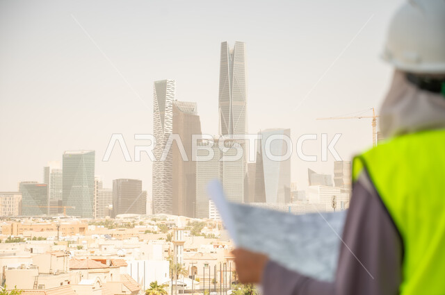  تطور القطاع الهندسي في المملكة، خلفية أبراج وناطحات سحاب، صورة خلفية لمهندسة معمارية عربية سعودية خليجية تحمل مخطط البناء وتتأمل معالم المدينة ، مهندسة انشاءات محجبة ترتدي بدلة السلامة في العمل، لباس رسمي للعمل ، خوذة العمل, مراجعة المخططات الهندسية وتدقيقها