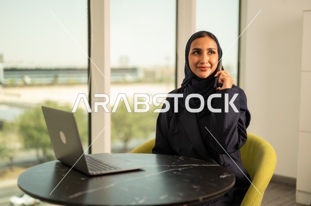 اجراء مكالمة هاتفية عبر الهاتف، ادارة الأعمال عن بعد، استخدام جهاز تقني حديث ومتطور، امرأة عربية خليجية سعودية ترتدي العباءة والحجاب تقوم باستخدام الحاسوب المحمول في المكتب، دمج التكنولوجيا بالعمل، إنجاز المهام المطلوبة عبر اللابتوب، مهنة ووظيفة نسائية مكتبية