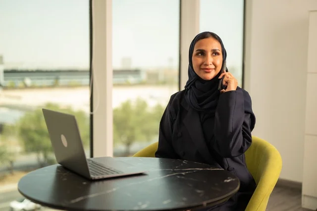 اجراء مكالمة هاتفية عبر الهاتف، ادارة الأعمال عن بعد، استخدام جهاز تقني حديث ومتطور، امرأة عربية خليجية سعودية ترتدي العباءة والحجاب تقوم باستخدام الحاسوب المحمول في المكتب، دمج التكنولوجيا بالعمل، إنجاز المهام المطلوبة عبر اللابتوب، مهنة ووظيفة نسائية مكتبية