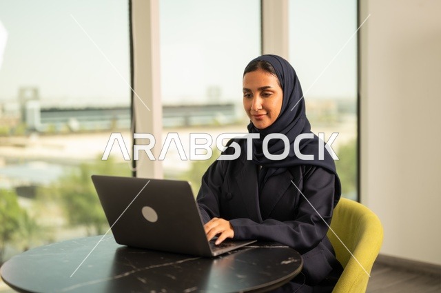 Remote business management, using a modern and advanced technical device, a Saudi Arabian Gulf woman wearing an abaya and hijab using a laptop in the office, integrating technology into work, completing required tasks via laptop, a female office profession and job