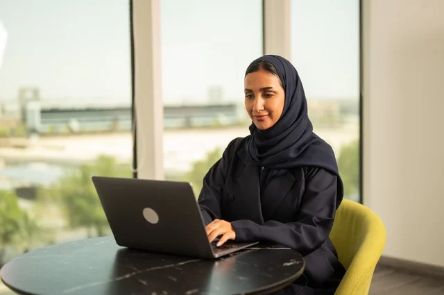 Remote business management, using a modern and advanced technical device, a Saudi Arabian Gulf woman wearing an abaya and hijab using a laptop in the office, integrating technology into work, completing required tasks via laptop, a female office profession and job
