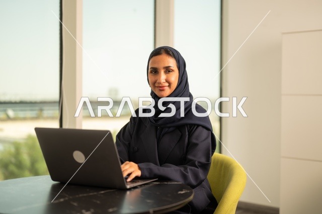 Remote business management, using a modern and advanced technical device, a Saudi Arabian Gulf woman wearing an abaya and hijab using a laptop in the office, integrating technology into work, completing required tasks via laptop, a female office profession and job