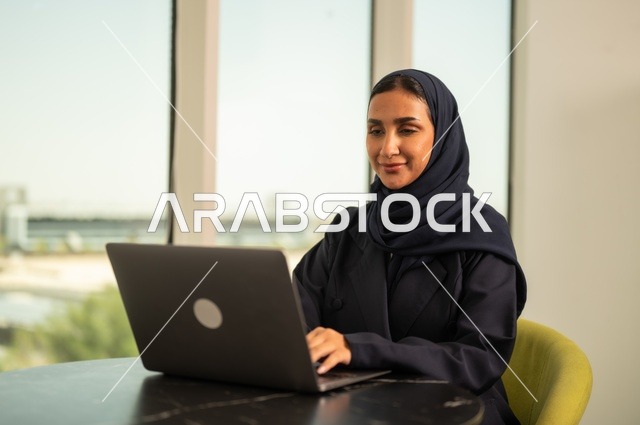 ادارة الأعمال عن بعد، استخدام جهاز تقني حديث ومتطور، امرأة عربية خليجية سعودية ترتدي العباءة والحجاب تقوم باستخدام الحاسوب المحمول في المكتب، دمج التكنولوجيا بالعمل، إنجاز المهام المطلوبة عبر اللابتوب، مهنة ووظيفة نسائية مكتبية