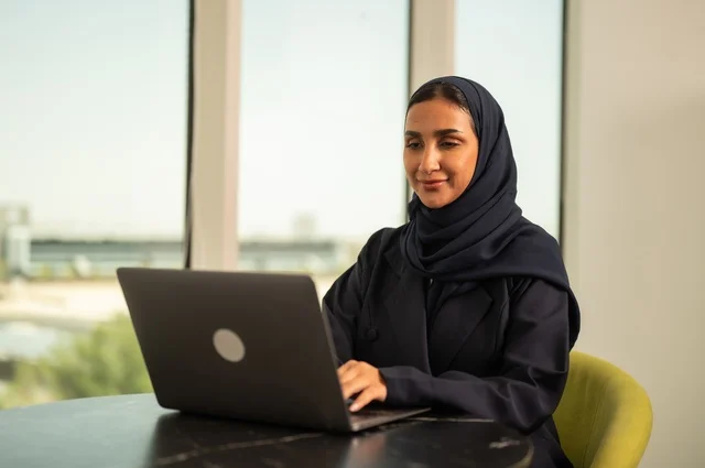 ادارة الأعمال عن بعد، استخدام جهاز تقني حديث ومتطور، امرأة عربية خليجية سعودية ترتدي العباءة والحجاب تقوم باستخدام الحاسوب المحمول في المكتب، دمج التكنولوجيا بالعمل، إنجاز المهام المطلوبة عبر اللابتوب، مهنة ووظيفة نسائية مكتبية