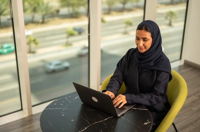 ادارة الأعمال عن بعد، استخدام جهاز تقني حديث ومتطور، امرأة عربية خليجية سعودية ترتدي العباءة والحجاب تقوم باستخدام الحاسوب المحمول في المكتب، دمج التكنولوجيا بالعمل، إنجاز المهام المطلوبة عبر اللابتوب، مهنة ووظيفة نسائية مكتبية