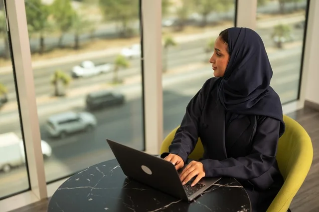 ادارة الأعمال عن بعد، استخدام جهاز تقني حديث ومتطور، امرأة عربية خليجية سعودية ترتدي العباءة والحجاب تقوم باستخدام الحاسوب المحمول في المكتب، دمج التكنولوجيا بالعمل، إنجاز المهام المطلوبة عبر اللابتوب، مهنة ووظيفة نسائية مكتبية