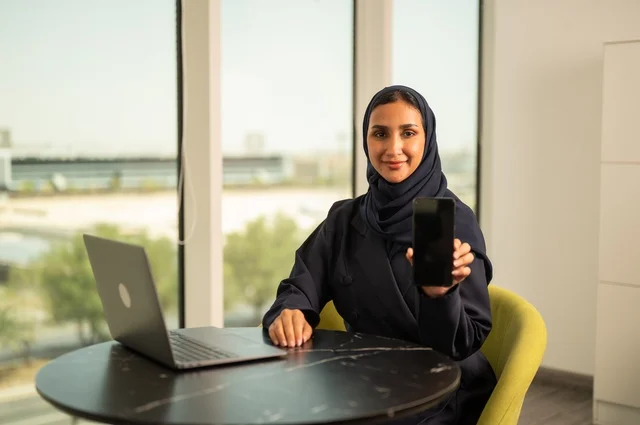 Blank black screen displayed on phone, remote business management, using a modern and advanced technical device, a Saudi Arabian Gulf woman wearing an abaya and hijab using a laptop in the office, integrating technology into work, completing required tasks via laptop, a female office profession and job