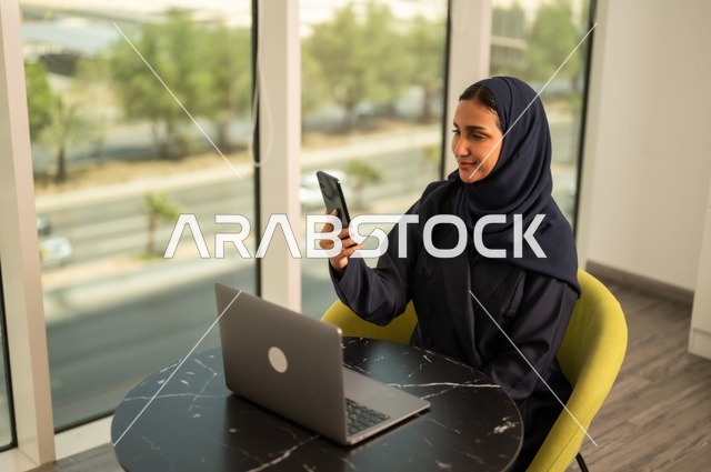 تصفح مواقع التواصل الاجتماعي عبر الهاتف، ادارة الأعمال عن بعد، استخدام جهاز تقني حديث ومتطور، امرأة عربية خليجية سعودية ترتدي العباءة والحجاب تقوم باستخدام الحاسوب المحمول في المكتب، دمج التكنولوجيا بالعمل، إنجاز المهام المطلوبة، مهنة ووظيفة نسائية مكتبية