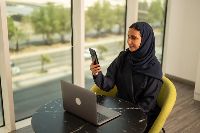 تصفح مواقع التواصل الاجتماعي عبر الهاتف، ادارة الأعمال عن بعد، استخدام جهاز تقني حديث ومتطور، امرأة عربية خليجية سعودية ترتدي العباءة والحجاب تقوم باستخدام الحاسوب المحمول في المكتب، دمج التكنولوجيا بالعمل، إنجاز المهام المطلوبة، مهنة ووظيفة نسائية مكتبية