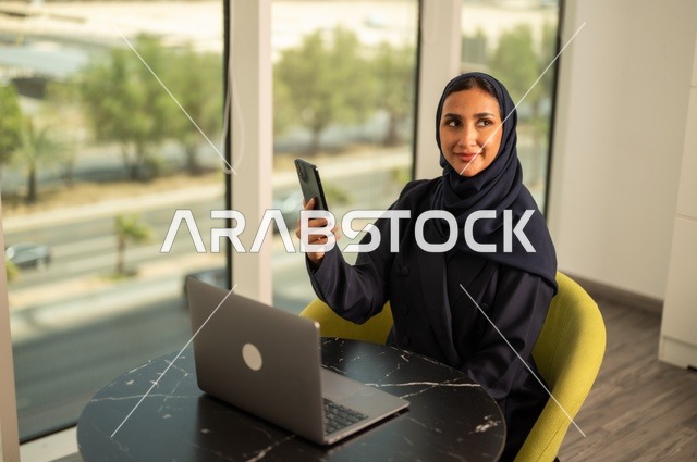 تصفح مواقع التواصل الاجتماعي عبر الهاتف، ادارة الأعمال عن بعد، استخدام جهاز تقني حديث ومتطور، امرأة عربية خليجية سعودية ترتدي العباءة والحجاب تقوم باستخدام الحاسوب المحمول في المكتب، دمج التكنولوجيا بالعمل، إنجاز المهام المطلوبة، مهنة ووظيفة نسائية مكتبية