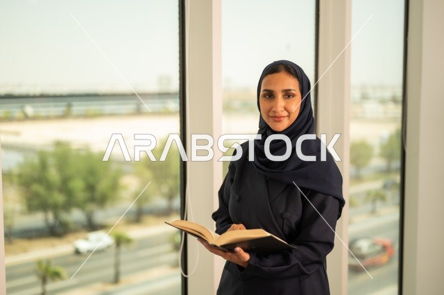 ممارسة هواية المطالعة, مفهوم الشغف بالقراءة, ثقافة التعلم الذاتي، قضاء الوقت بالتثقف، الاستمتاع بقراءة الكتب والقصص والروايات، امرأة عربية خليجية سعودية محجبة ترتدي العباءة تقف في المكتب تحمل بيديها كتاب، تنمية وتعزيز المعلومات العامة