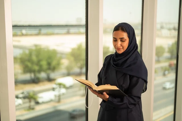 ممارسة هواية المطالعة, مفهوم الشغف بالقراءة, ثقافة التعلم الذاتي، قضاء الوقت بالتثقف، الاستمتاع بقراءة الكتب والقصص والروايات، امرأة عربية خليجية سعودية محجبة ترتدي العباءة تقف في المكتب تحمل بيديها كتاب، تنمية وتعزيز المعلومات العامة