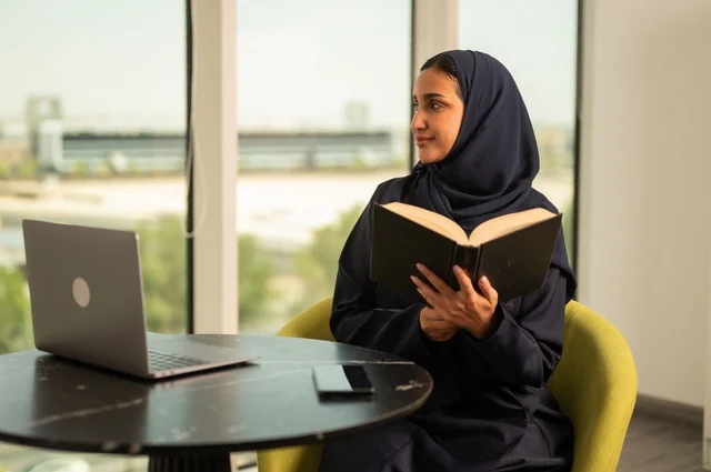 Practicing the hobby of reading, the concept of passion for reading, remote business management, using a modern and advanced technical device, a Saudi Arabian Gulf woman wearing an abaya and hijab using a laptop in the office, integrating technology into work, completing required tasks, a female office profession and job.