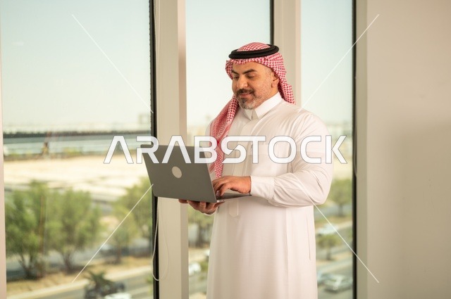 الشركات والمكاتب في المملكة العربية السعودية، إدارة شؤون الأعمال وتنظيمها، استخدام جهاز تقني حديث ومتطور، مهنة ووظيفة ادارية مكتبية، موظف عربي خليجي سعودي يرتدي الثوب التقليدي يقوم بالعمل على الحاسوب المحمول داخل المكتب، خلفية أبراج الرياض