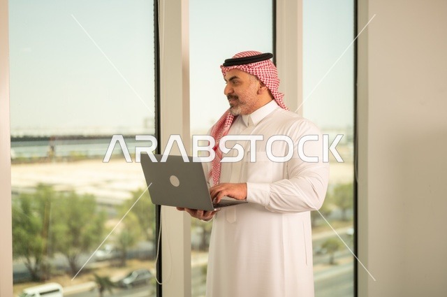 الشركات والمكاتب في المملكة العربية السعودية، إدارة شؤون الأعمال وتنظيمها، استخدام جهاز تقني حديث ومتطور، مهنة ووظيفة ادارية مكتبية، موظف عربي خليجي سعودي يرتدي الثوب التقليدي يقوم بالعمل على الحاسوب المحمول داخل المكتب، خلفية أبراج الرياض
