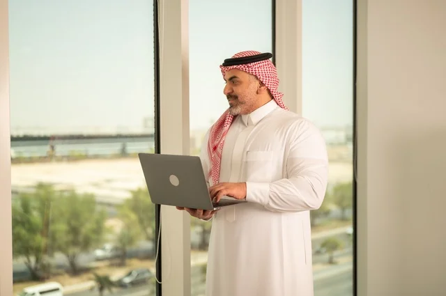 الشركات والمكاتب في المملكة العربية السعودية، إدارة شؤون الأعمال وتنظيمها، استخدام جهاز تقني حديث ومتطور، مهنة ووظيفة ادارية مكتبية، موظف عربي خليجي سعودي يرتدي الثوب التقليدي يقوم بالعمل على الحاسوب المحمول داخل المكتب، خلفية أبراج الرياض