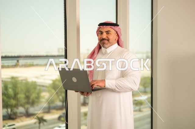 الشركات والمكاتب في المملكة العربية السعودية، إدارة شؤون الأعمال وتنظيمها، استخدام جهاز تقني حديث ومتطور، مهنة ووظيفة ادارية مكتبية، موظف عربي خليجي سعودي يرتدي الثوب التقليدي يقوم بالعمل على الحاسوب المحمول داخل المكتب، خلفية أبراج الرياض