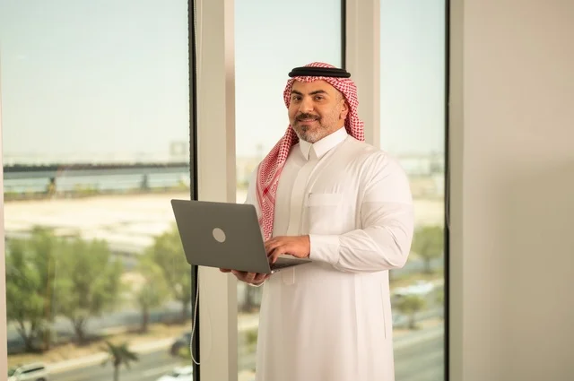 الشركات والمكاتب في المملكة العربية السعودية، إدارة شؤون الأعمال وتنظيمها، استخدام جهاز تقني حديث ومتطور، مهنة ووظيفة ادارية مكتبية، موظف عربي خليجي سعودي يرتدي الثوب التقليدي يقوم بالعمل على الحاسوب المحمول داخل المكتب، خلفية أبراج الرياض
