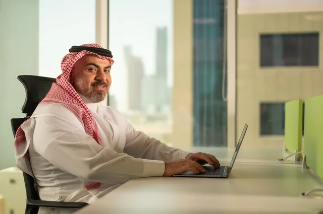 إدارة شؤون الأعمال وتنظيمها، استخدام جهاز تقني حديث ومتطور، مهنة ووظيفة ادارية مكتبية، موظف عربي خليجي سعودي يرتدي الثوب التقليدي يقوم بالعمل على الحاسوب المحمول داخل المكتب، الشركات والمكاتب في المملكة العربية السعودية، خلفية أبراج الرياض