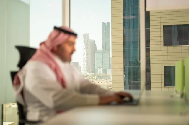 إدارة شؤون الأعمال وتنظيمها، استخدام جهاز تقني حديث ومتطور، مهنة ووظيفة ادارية مكتبية، موظف عربي خليجي سعودي يرتدي الثوب التقليدي يقوم بالعمل على الحاسوب المحمول داخل المكتب، الشركات والمكاتب في المملكة العربية السعودية، خلفية أبراج الرياض