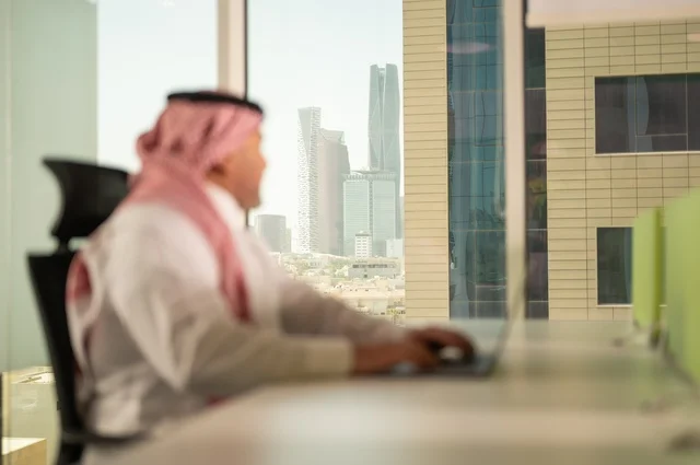 إدارة شؤون الأعمال وتنظيمها، استخدام جهاز تقني حديث ومتطور، مهنة ووظيفة ادارية مكتبية، موظف عربي خليجي سعودي يرتدي الثوب التقليدي يقوم بالعمل على الحاسوب المحمول داخل المكتب، الشركات والمكاتب في المملكة العربية السعودية، خلفية أبراج الرياض