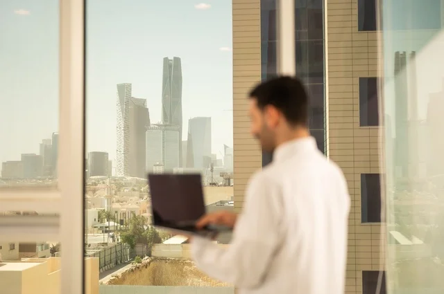 Using a modern and advanced technical device, an office administrative profession and job, a Saudi Arabian Gulf Arab employee wearing traditional clothing working on a laptop inside the office, companies and offices in the Kingdom of Saudi Arabia, managing and organizing business affairs, Riyadh Towers background