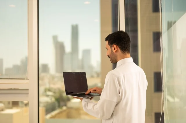 استخدام جهاز تقني حديث ومتطور، مهنة ووظيفة ادارية مكتبية، موظف عربي خليجي سعودي يرتدي الثوب التقليدي يقوم بالعمل على الحاسوب المحمول داخل المكتب، الشركات والمكاتب في المملكة العربية السعودية، إدارة شؤون الأعمال وتنظيمها، خلفية أبراج الرياض