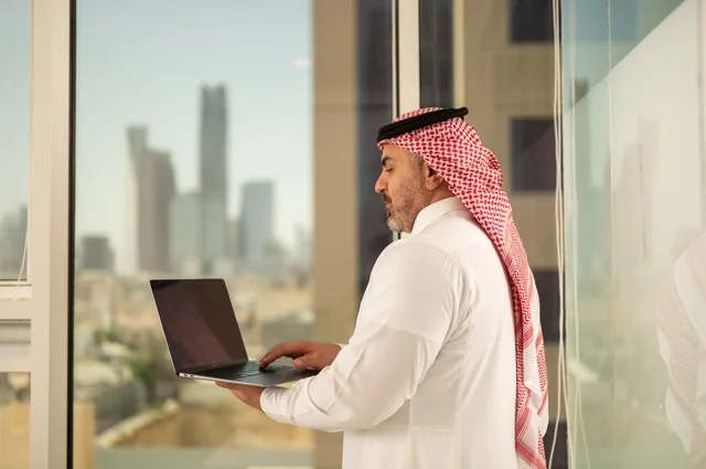 استخدام جهاز تقني حديث ومتطور، مهنة ووظيفة ادارية مكتبية، موظف عربي خليجي سعودي يرتدي الثوب التقليدي يقوم بالعمل على الحاسوب المحمول داخل المكتب، الشركات والمكاتب في المملكة العربية السعودية، إدارة شؤون الأعمال وتنظيمها، خلفية أبراج الرياض