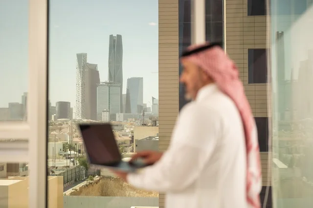 استخدام جهاز تقني حديث ومتطور، مهنة ووظيفة ادارية مكتبية، موظف عربي خليجي سعودي يرتدي الثوب التقليدي يقوم بالعمل على الحاسوب المحمول داخل المكتب، الشركات والمكاتب في المملكة العربية السعودية، إدارة شؤون الأعمال وتنظيمها، خلفية أبراج الرياض