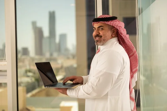 استخدام جهاز تقني حديث ومتطور، مهنة ووظيفة ادارية مكتبية، موظف عربي خليجي سعودي يرتدي الثوب التقليدي يقوم بالعمل على الحاسوب المحمول داخل المكتب، الشركات والمكاتب في المملكة العربية السعودية، إدارة شؤون الأعمال وتنظيمها، خلفية أبراج الرياض