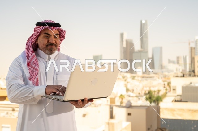 الشركات والمكاتب في المملكة العربية السعودية، إدارة شؤون الأعمال وتنظيمها، استخدام جهاز تقني حديث ومتطور، مهنة ووظيفة ادارية مكتبية، موظف عربي خليجي سعودي يرتدي الثوب التقليدي يقوم بالعمل على الحاسوب المحمول داخل المكتب، خلفية أبراج الرياض