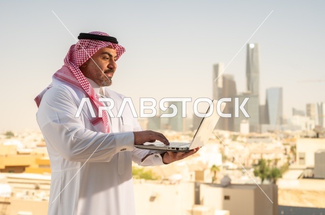 الشركات والمكاتب في المملكة العربية السعودية، إدارة شؤون الأعمال وتنظيمها، استخدام جهاز تقني حديث ومتطور، مهنة ووظيفة ادارية مكتبية، موظف عربي خليجي سعودي يرتدي الثوب التقليدي يقوم بالعمل على الحاسوب المحمول داخل المكتب، خلفية أبراج الرياض