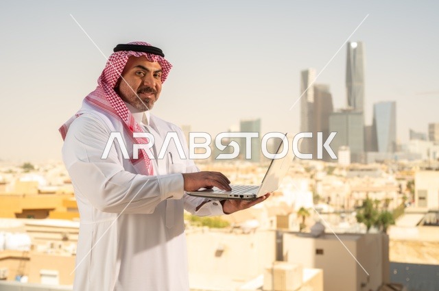 Companies and offices in the Kingdom of Saudi Arabia, managing and organizing business affairs, using modern and advanced technical equipment, an administrative office profession and job, a Saudi Gulf Arab employee wearing traditional clothing working on a laptop inside the office, Riyadh towers background