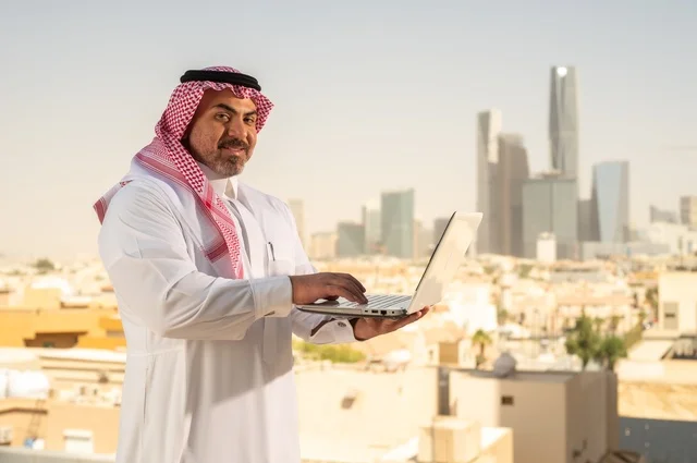 الشركات والمكاتب في المملكة العربية السعودية، إدارة شؤون الأعمال وتنظيمها، استخدام جهاز تقني حديث ومتطور، مهنة ووظيفة ادارية مكتبية، موظف عربي خليجي سعودي يرتدي الثوب التقليدي يقوم بالعمل على الحاسوب المحمول داخل المكتب، خلفية أبراج الرياض