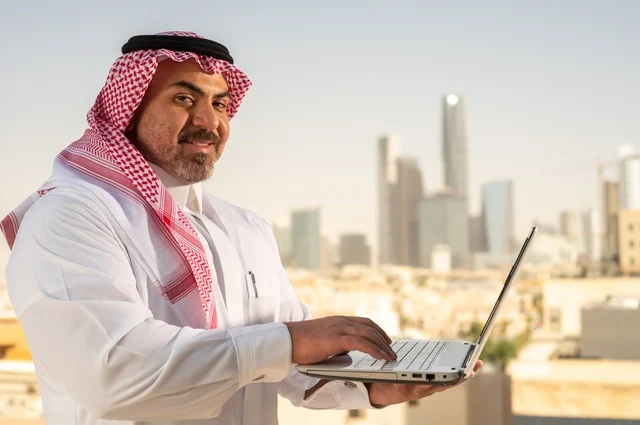 مهنة ووظيفة ادارية مكتبية، استخدام جهاز تقني حديث ومتطور، موظف عربي خليجي سعودي يرتدي الثوب التقليدي يقوم بالعمل على الحاسوب المحمول داخل المكتب، الشركات والمكاتب في المملكة العربية السعودية، إدارة شؤون الأعمال وتنظيمها، خلفية أبراج الرياض