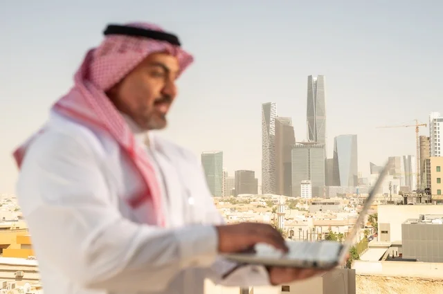 مهنة ووظيفة ادارية مكتبية، استخدام جهاز تقني حديث ومتطور، موظف عربي خليجي سعودي يرتدي الثوب التقليدي يقوم بالعمل على الحاسوب المحمول داخل المكتب، الشركات والمكاتب في المملكة العربية السعودية، إدارة شؤون الأعمال وتنظيمها، خلفية أبراج الرياض