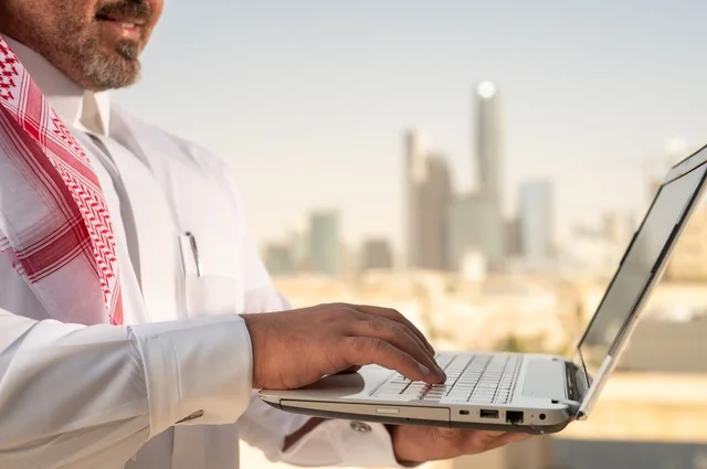 Administrative office profession and job, using modern and advanced technical equipment, a Saudi Arabian Gulf Arab employee wearing traditional clothing working on a laptop inside the office, companies and offices in the Kingdom of Saudi Arabia, managing and organizing business affairs, Riyadh towers background