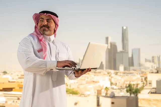 مهنة ووظيفة ادارية مكتبية، استخدام جهاز تقني حديث ومتطور، موظف عربي خليجي سعودي يرتدي الثوب التقليدي يقوم بالعمل على الحاسوب المحمول داخل المكتب، الشركات والمكاتب في المملكة العربية السعودية، إدارة شؤون الأعمال وتنظيمها، خلفية أبراج الرياض