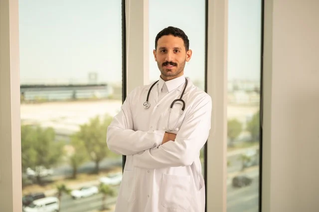 العمل في القطاع الصحي، مفهوم الفحص الطبي، تقديم الخدمات العلاجية للمرضى, طبيب عربي خليجي سعودي يرتدي الثوب التقليدي والمعطف الطبي يضع السماعة الطبية يقف بأيدي متقاطعة يعمل في المراكز الصحية والمستشفيات، خلفية أبراج الرياض