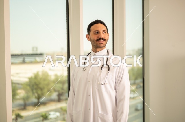 مفهوم الفحص الطبي، العمل في القطاع الصحي، تقديم الخدمات العلاجية للمرضى, طبيب عربي خليجي سعودي يرتدي الثوب التقليدي والمعطف الطبي يضع السماعة الطبية يعمل في المراكز الصحية والمستشفيات، خلفية أبراج الرياض