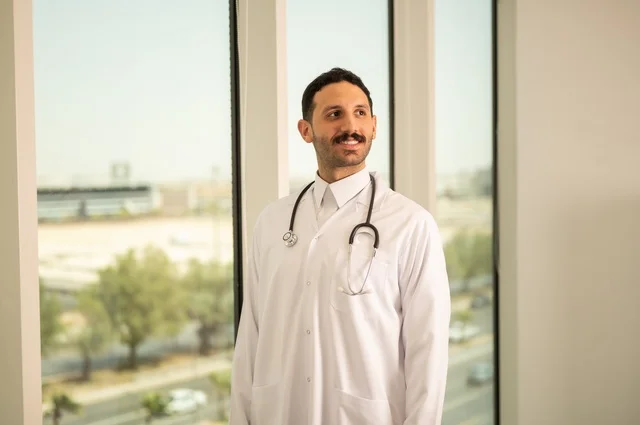 مفهوم الفحص الطبي، العمل في القطاع الصحي، تقديم الخدمات العلاجية للمرضى, طبيب عربي خليجي سعودي يرتدي الثوب التقليدي والمعطف الطبي يضع السماعة الطبية يعمل في المراكز الصحية والمستشفيات، خلفية أبراج الرياض