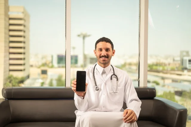 Blank black screen view on mobile phone, medical examination concept, working in healthcare sector, providing medical services to patients, Saudi Arabian Gulf doctor wearing traditional thobe and medical coat wearing stethoscope working in healthcare centers and hospitals, Riyadh towers background