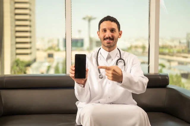 عرض شاشة سوداء فارغة عبر الهاتف المحمول، مفهوم الفحص الطبي، العمل في القطاع الصحي، تقديم الخدمات العلاجية للمرضى, طبيب عربي خليجي سعودي يرتدي الثوب التقليدي والمعطف الطبي يضع السماعة الطبية يعمل في المراكز الصحية والمستشفيات، خلفية أبراج الرياض