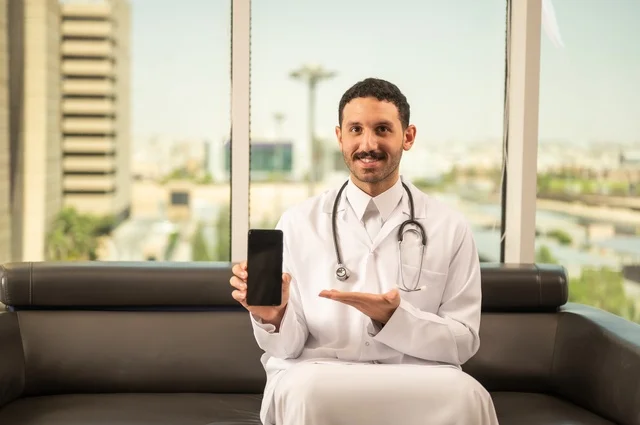 عرض شاشة سوداء فارغة عبر الهاتف المحمول، مفهوم الفحص الطبي، العمل في القطاع الصحي، تقديم الخدمات العلاجية للمرضى, طبيب عربي خليجي سعودي يرتدي الثوب التقليدي والمعطف الطبي يضع السماعة الطبية يعمل في المراكز الصحية والمستشفيات، خلفية أبراج الرياض